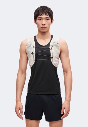 Joven con chaleco de hidratación beige sobre una camiseta deportiva negra sin mangas y pantalones cortos negros, de pie frente a un fondo claro y liso.