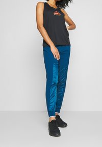 Schwarzes ärmelloses Top mit Nike Trail-Logo, kombiniert mit blauen Hosen mit glänzenden Seitenpaneelen und schwarzen Sportschuhen.