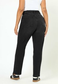 Mujer que lleva unos vaqueros negros de talle alto y piernas rectas, camiseta blanca, zapatillas negras con rayas blancas, de pie delante de un fondo claro y liso.