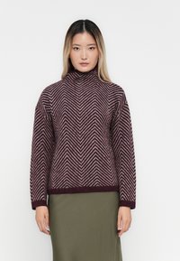 Woman wearing a high-neck, long-sleeve sweater with a maroon and white chevron pattern and an olive green skirt, standing against a white background.