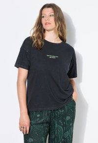 Sort bomulds t-shirt med korte ærmer og en afslappet pasform, med grøn tekst på fronten. Modellen har mønstrede grønne bukser på.