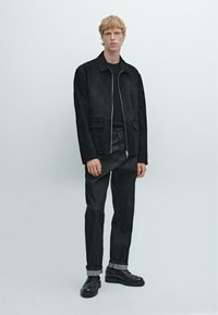 Giacca in suede nera con chiusura a zip, due tasche frontali; abbinata a jeans in denim scuro e scarpe nere stringate con risvolti.