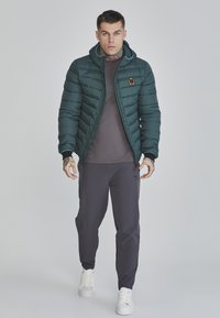 Mörkgrön puffjacka med huva, som har ett quiltat mönster och en liten emblem. Matchad med grå byxor och vita sneakers.