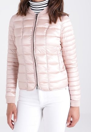 Kvinde iført lyserød quiltet jakke med frontlynlås over sort-hvid stribet rullekrave og hvide bukser, stående med afslappede hænder.