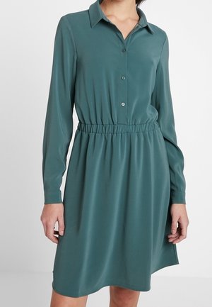 Langärmliges türkisfarbenes Hemdkleid mit Kragen, Knopfleiste vorne, elastischer Taille und knielangem Saum, getragen von einer stehenden Person.