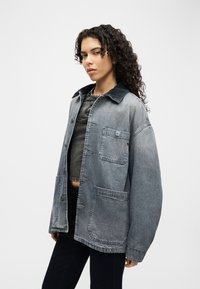 Jeune femme aux cheveux bouclés portant une veste en jean délavée et oversize par-dessus un top court foncé et un pantalon noir, debout devant un fond uni.