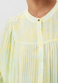 Camicia button-up gialla limone con design plissettato, tessuto leggero, colletto rotondo e motivo di foglie azzurre morbide. Presenta piccoli bottoni.