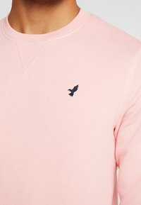 Zbliżenie osoby noszącej jasnoróżową bluzę z okrągłym dekoltem, z małym, ciemnym haftowanym logo ptaka na piersi.