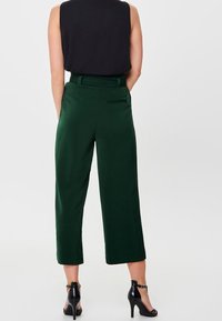 Pantaloni verdi a cropped con una texture liscia, vita alta e tasche laterali, abbinati a un top senza maniche nero e scarpe con tacco.