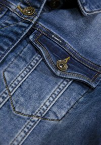 Denim stof in medium blauw, met gouden stiksels, een enkele borstzak met een knoopsluiting en een label met de tekst "Garcia."