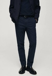 Pantaloni blu con un tessuto testurizzato, design affusolato, tasche laterali, abbinati a un blazer blu coordinato e stivali neri lucidi.