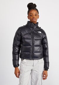 Črn puhasti plašč z sijočo površino, z visokim ovratnikom, sprednjim zadrgo in logotipom North Face na levem prsnem košu. Tekstuirana, prešita zasnova.