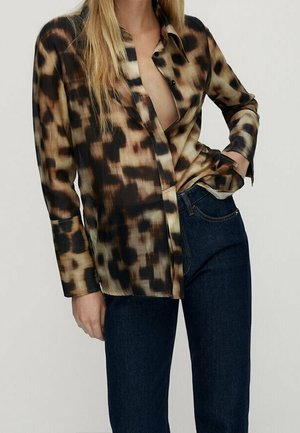 Femme portant une chemise à manches longues marron et noire à motifs abstraits, partiellement déboutonnée, avec un jean bleu foncé, une main dans la poche.