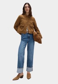 Bruine suède jas met een kraag en twee voorzakken, gecombineerd met blauwe opgerolde jeans en tan instapschoenen. Geaccentueerd door zichtbare knopen.