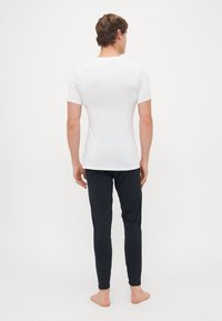 Homme debout pieds nus, de dos, portant une chemise blanche ajustée à manches courtes et un pantalon noir slim, sur un fond uni.