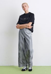 Zwarte oversized t-shirt met reflecterende tekst, gecombineerd met glanzende grijze wijde broek met witte strepen en accentpatronen. Zwarte sneakers.