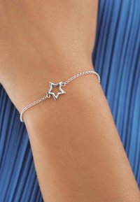 Pulsera de plata con un colgante en forma de estrella, que presenta una cadena delicada y un acabado pulido. La pulsera reposa sobre una muñeca tostada.
