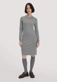 Donna con capelli bob scuri che indossa un abito a maglia lungo grigio, calze grigie fino al ginocchio e mocassini con frange marroni scuri, in piedi su uno sfondo chiaro.