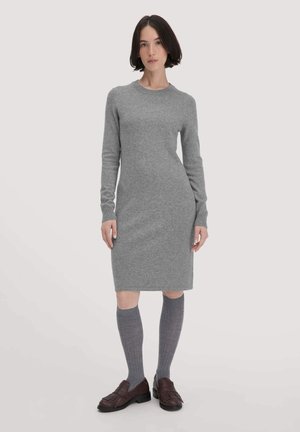 Vrouw met donker bobhaar, gekleed in een grijs lange mouw gebreide jurk, kniehoog grijze sokken en donkerbruine franjesloafers, staand op een lichte achtergrond.