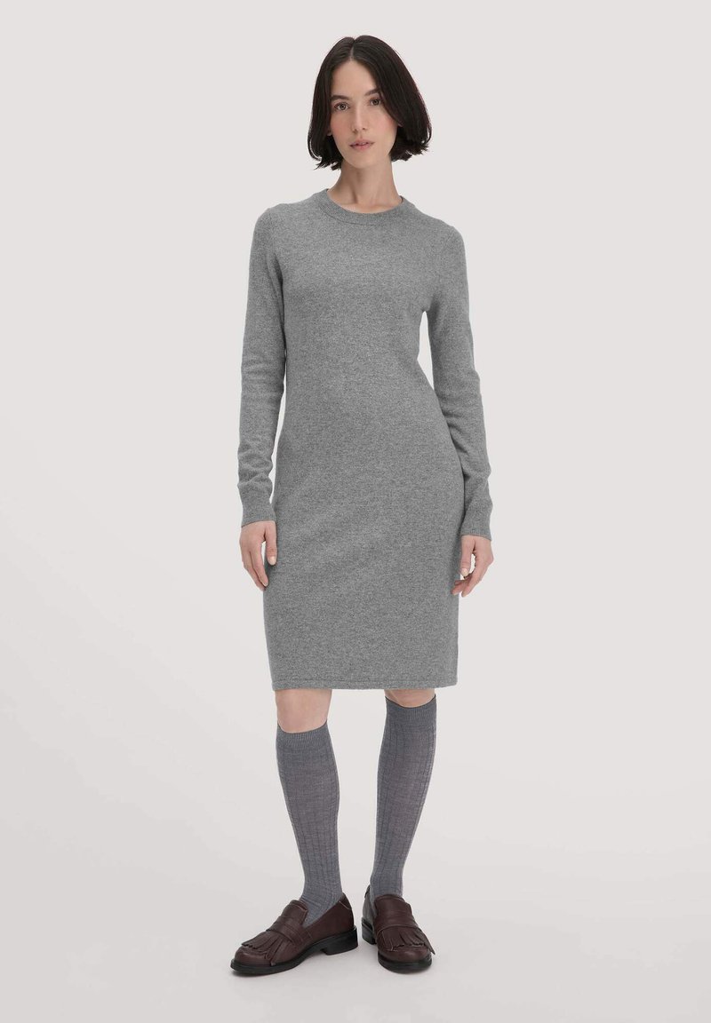 Donna con capelli bob scuri che indossa un abito a maglia lungo grigio, calze grigie fino al ginocchio e mocassini con frange marroni scuri, in piedi su uno sfondo chiaro.