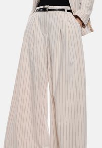 Pantaloni a gamba larga in tessuto beige chiaro con righe verticali scure, caratterizzati da pieghe, vita alta e una cintura nera.