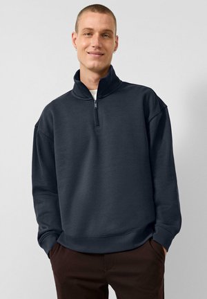 Jonge man met kort haar die een donkergrijze quarter-zip trui en een bruine broek draagt, staat met handen in de zakken en glimlacht.