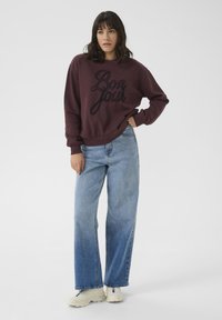 Weinroter Sweatshirt mit "Bon Jour" Stickerei, kombiniert mit hellblauen, weit geschnittenen Jeans und weißen Sneakers. Schlichte, lässige Passform.