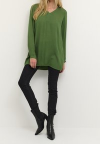 Groen shirt met lange mouwen, een v-hals en een rechte snit, gecombineerd met nauwsluitende zwarte spijkerbroek en zwarte puntige enkellaarzen. Eenvoudig ontwerp.