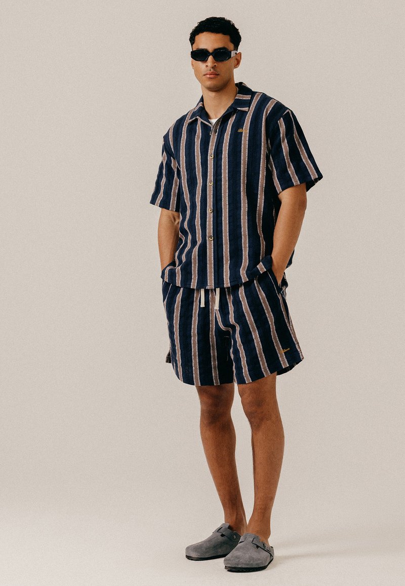 Homme portant une chemise à manches courtes rayée verticalement bleu marine et beige et un short assorti, des lunettes de soleil noires et des chaussures grises sans lacets, debout les mains dans les poches.