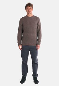 Suéter marrón de punto acanalado con cuello redondo, combinado con pantalones gris oscuro y zapatillas negras, de pie contra un fondo blanco.