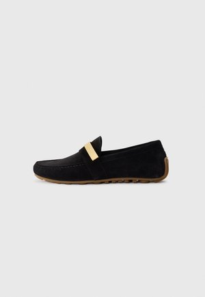 Mocassin en suede noir avec une bande en métal doré sur le devant, semelle plate en caoutchouc avec grip texturé, et un design à bout rond.