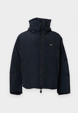 Jeune homme portant une veste à capuche noire, un t-shirt blanc graphique, un jean bleu clair, des baskets blanches et une casquette bleu marine, debout devant un fond blanc.