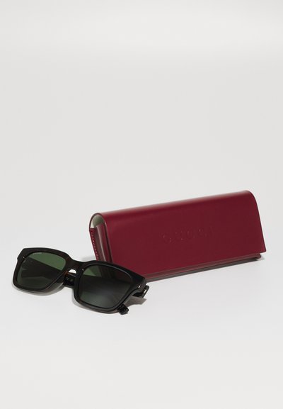 Schwarz gerahmte Sonnenbrille mit quadratischem Rahmen und dunkelgrünen Gläsern neben einem geschlossenen roten Leder-Etui auf weißem Hintergrund.