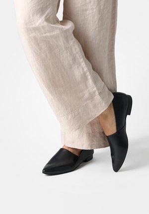 Schwarze flache Schuhe aus Leder mit spitzer Zehenpartie, glatter Oberfläche und niedrigem Absatz, kombiniert mit hellen, locker sitzenden Hosen.