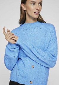 Svetlo modri pleteni kardigan z rebrastim ovratnikom, prevelikimi rokavi in vzorčenimi očesnimi detajli. Sprednje zapiranje z gumbi in sproščen kroj.