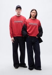 Κόκκινες φούτερ με το λογότυπο "tommy", συνδυασμένες με σκουρόχρωμα τζιν. Ο ανδρικός μοντέλο φοράει μαύρο καπέλο, η γυναικεία μοντέλο έχει μαύρο μπουφάν.