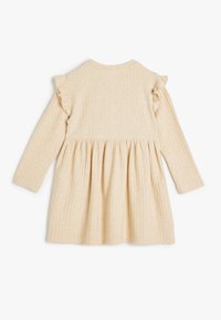 Kroczona, długa sukienka z długimi rękawami w jasnym beżu, z falbanami na ramionach i dopasowanym fasonem, który rozszerza się w żakardową spódnicę.