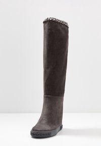 Botte haute en daim gris foncé montant jusqu'au genou, avec un détail en chaîne sur le bord supérieur et une semelle en caoutchouc texturée sur fond blanc uni.