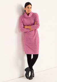 Rosa tröjklänning med en båtformad halsringning, långa ärmar, sidodragkedjor och ett strukturerat stickat mönster. Matchas med svarta leggings och ankelstövlar.