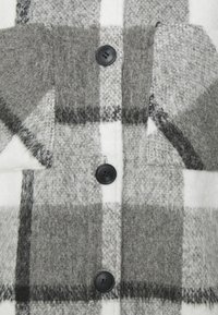 Cappotto in misto lana con motivo a scacchi grigio e bianco. Quattro bottoni neri sul davanti e due tasche sul petto. Texture morbida.