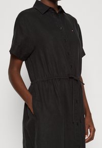 Černé košilové šaty s krátkými rukávy, vyrobené z lehké tkaniny. Mají zúžený pas a boční kapsy pro praktičnost.