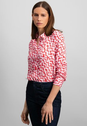 Red and pink zigzag patterned button-up shirt, long sleeves, collared, paired with dark blue jeans. Fine texture, casual design.