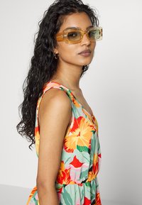 Vestido sem mangas com estampas florais vibrantes em laranja, rosa e verde; usado com óculos de sol retangulares amarelos e brincos de argola.