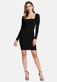 Czarna sukienka z prążkowanej tkaniny, z długimi rękawami i prostokątnym dekoltem, dopasowana sylwetka, długość do połowy uda, w zestawie z czarnymi sandałami na obcasie.