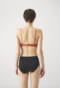 Bra with thin straps in rust color and hook closure, paired with high-waisted black brief featuring scalloped edges. Back view.