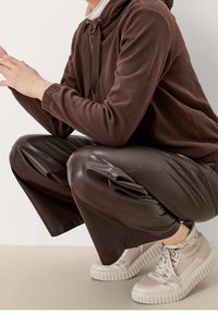 Brun fleecehoodie med dragsko, tillsammans med mörkbruna veckade fusk läderbyxor och ljusa klumpiga sneakers.