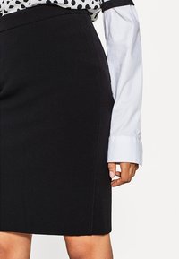 Zwarte potloodrok in een gladde stof, met een getailleerd silhouet en subtiele naden. Gecombineerd met een witte blouse met lange mouwen.