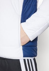 Giacca a vento imbottita con corpo bianco e accenti blu marine, tasca laterale con zip, texture trapuntata e polsini elasticizzati. Adatta per un abbigliamento casual.