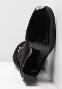 Botín ancho de ante negro con un tacón brillante negro y suela texturizada. Un detalle de cremallera lateral es visible a lo largo del eje.