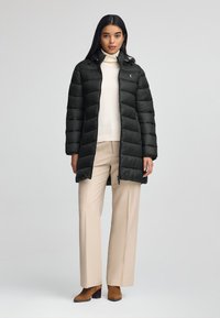 Czarna puchowa kurtka z poziomym pikowaniem, wysokim kołnierzem i zamkiem z przodu. Noszona na kremowym swetrze i beżowych spodniach, w zestawie z brązowymi botkami do kostek.